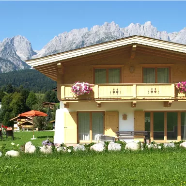 A charming alpine house with a balcony and flower boxes. In the background, majestic mountains rise beneath a clear blue sky.