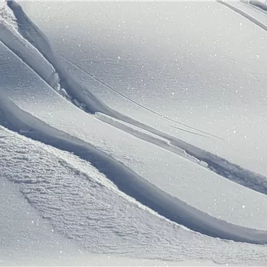 Ein sanfter Schnee-Hügel mit zarten Wellen und glitzernden Kristallen. Die weißen Linien zeigen die Spuren im frischen Schnee.