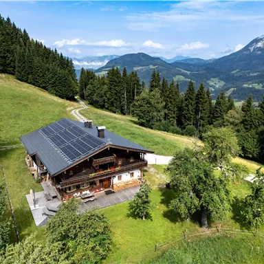 Ein charmantes Haus in den Bergen umgeben von grüner Natur und hohen Bäumen. Der Himmel ist klar und es ist eine ruhige, einladende Landschaft.