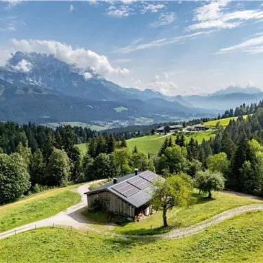 Eine malerische Landschaft mit einem Holzhäuschen auf einer Wiese. Im Hintergrund sind beeindruckende Berge und ein klarer Himmel zu sehen.