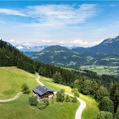 Eine malerische Landschaft mit grünen Wiesen und bewaldeten Bergen. Im Vordergrund steht ein gemütliches Haus am Fuß eines Hügels.