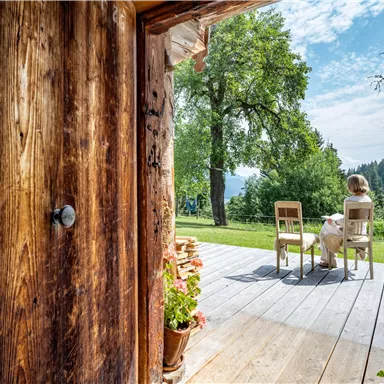 Eine offene Holztür zeigt eine Terrasse mit zwei Stühlen und einer Person, die den Ausblick genießt. Im Hintergrund sind grüne Bäume und ein klarer Himmel zu sehen.