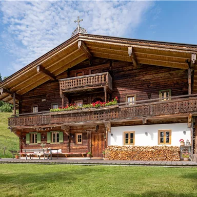 Ein traditionelles Holzhaus im alpinen Stil mit einem Balkon und bunten Blumen. Umgeben von grünen Wiesen und Bäumen unter einem klaren blauen Himmel.