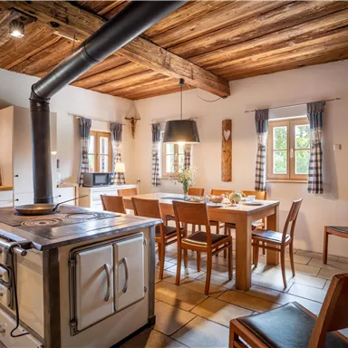 Eine gemütliche Küche mit rustikalem Design und Holzbalkendecke. Ein großer Esstisch steht im Mittelpunkt, umgeben von Stühlen und Fenster mit Vorhängen.
