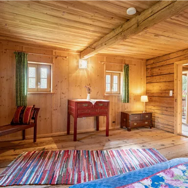 Ein gemütliches Holzzimmer mit buntem Teppich und natürlichem Licht. Es gibt einen kleinen Tisch, Stühle und Zugang zur Außentür.