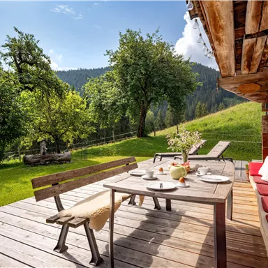 Eine gemütliche Terrasse mit einem Holztisch und Bänken, umgeben von grünen Bäumen und einer malerischen Berglandschaft. Die Sonne scheint und lädt zum Entspannen im Freien ein.