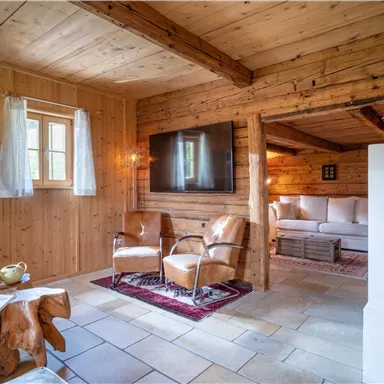 Ein gemütlicher Wohnraum mit Holzwänden und viel natürlichem Licht. Zwei Ledersessel stehen vor einem Fernseher und einem Sichtbereich mit einer gemütlichen Couch.