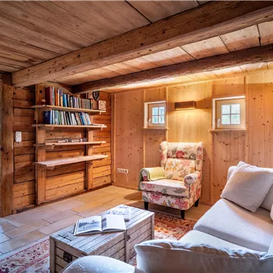 Ein gemütliches, rustikales Wohnzimmer mit Holzwänden und einem bequemen Sessel. Regale mit Büchern und sanftem Licht schaffen eine einladende Atmosphäre.
