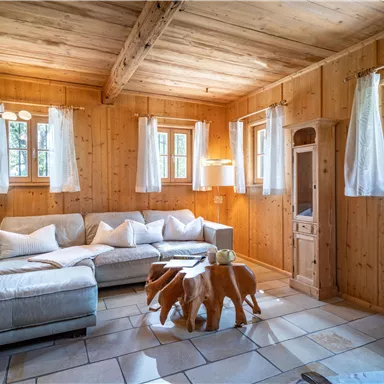 Ein gemütlicher Wohnraum mit Holzwänden und großen Fenstern. Die Einrichtung umfasst ein graues Sofa, einen modernen Couchtisch und einen angenehmen Lichteinfall.