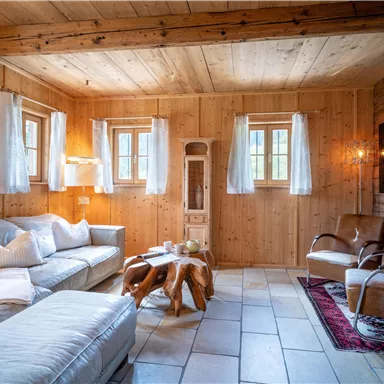 Ein gemütliches Wohnzimmer mit Holzverkleidung und große Fenster. Der Raum ist mit einem hellen Sofa und zwei Sesseln eingerichtet.