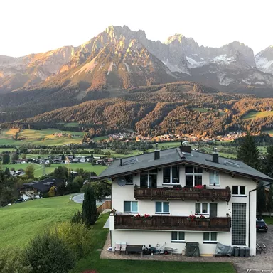 A beautiful house with balconies, surrounded by green meadows and mountains. The sun rays illuminate the landscape and create a picturesque atmosphere.