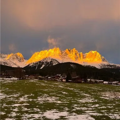 An impressive mountain landscape with golden light on the peaks. In the foreground, there is a green meadow with snow.
