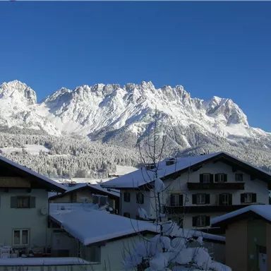 A snow-covered mountain landscape with high peaks and a clear blue sky. In the foreground, typical alpine houses can be seen.