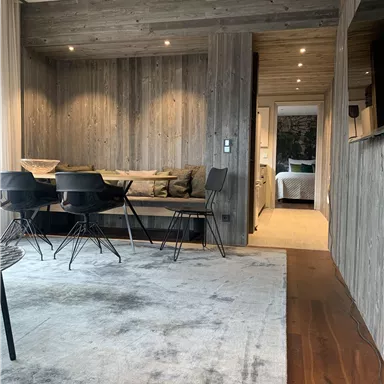 Ein moderner Wohnraum mit Holzvertäfelung und einer gemütlichen Sitzecke. Im Hintergrund ist ein minimalistisch gestalteter Schlafbereich zu sehen.