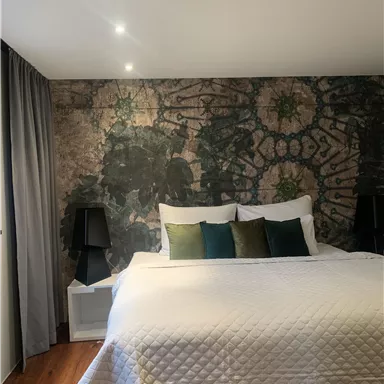 Ein stilvolles Schlafzimmer mit einem großen Bett und dekorativen Kissen. Die Wand ist mit einem auffälligen Muster versehen, und es gibt zwei Nachttische mit Lampen.