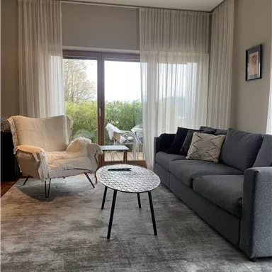 Ein gemütlicher Wohnraum mit einem grauen Sofa und einem weißen Sessel. Große Fenster lassen viel Licht herein und bieten Blick ins Grüne.