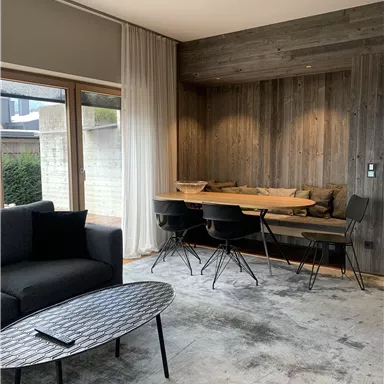 Ein modernes Wohnzimmer mit einem schwarzen Sofa und einem runden Tisch. Im Hintergrund befindet sich ein Essbereich aus Holz mit Stühlen.