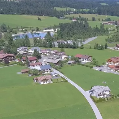 A picturesque landscape with a small village, surrounded by green meadows and forests. In the background, several buildings and a swimming pond can be seen.