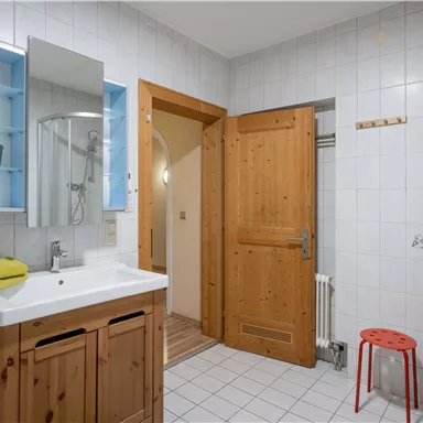 A modern bathroom with wooden details and a light tile wall. The furnishings include a sink, a mirror, and a shower.
