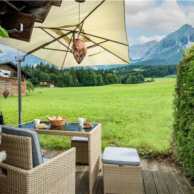 A cozy outdoor area with a table and chairs under a sunshade. In the background, green meadows and majestic mountains stretch out.