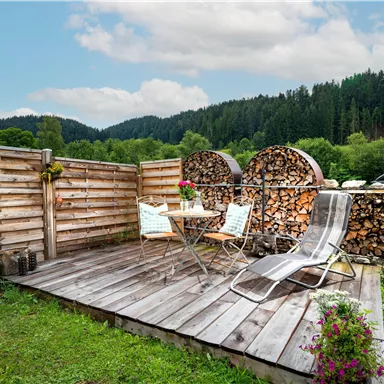A cozy terrace with a pile of wood in the background and a table with chairs. The surroundings are green with trees and a clear sky.