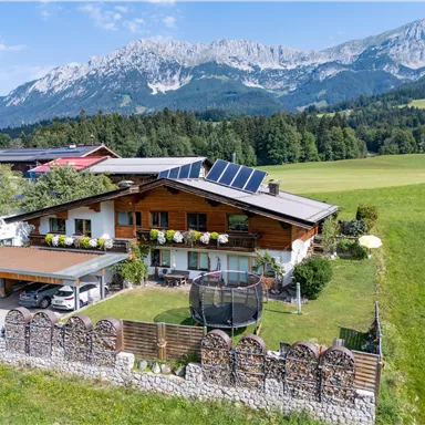 A cozy country house with solar panels in a picturesque landscape. In the background, high mountains and a green field can be seen.