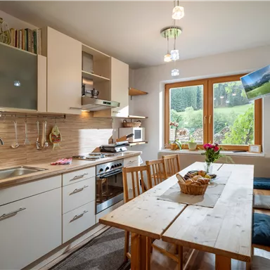 A modern kitchen with wooden shelves and a large table. A green landscape can be seen through the window.
