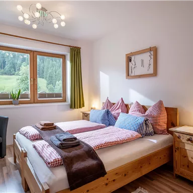 A cozy bedroom with a double bed and checkered pillows. Large windows offer a view of the green landscape.
