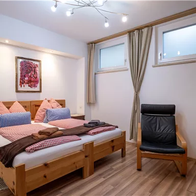 A cozy bedroom with a double bed, wooden furniture, and a swivel chair. The room has soft colors and large windows that provide natural light.