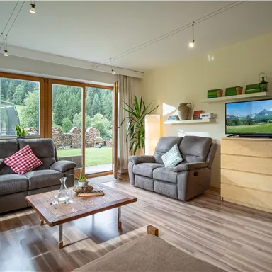 A cozy living room with modern furniture and large windows that offer a view of the garden. The television and the plants complement the inviting atmosphere.