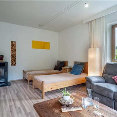 A cozy living room with a gray sofa and two wooden loungers. Large windows provide a view of the outside, and there are some decorative plants.