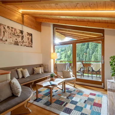 A cozy living room with modern wood design and large windows. The room is brightly lit and offers a view of a green-covered landscape.