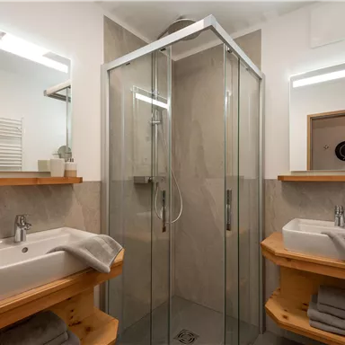 A modern bathroom with a glass shower and two sinks. Wooden shelves and plants create a cozy atmosphere.