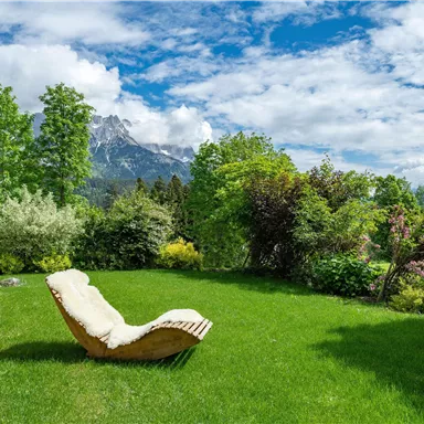 A beautiful garden with lush green grass and colorful plants. In the background, impressive mountains and a blue sky with clouds can be seen.