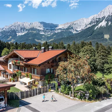 A traditional Alpine house surrounded by trees and meadows. In the background, impressive mountains rise under a blue sky.