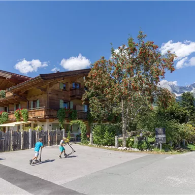 A charming wooden building is located in a picturesque landscape. Two children are riding their scooters along a road in front of the building.
