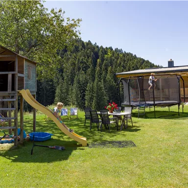 A play area with a climbing frame, a slide, and a trampoline. In the background, there are trees and a beautiful house.