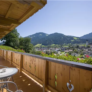 Ein Balkon mit einem Tisch und Stühlen, der eine wunderschöne Aussicht auf grüne Berge und ein Dorf bietet. Der Himmel ist klar und sonnig.