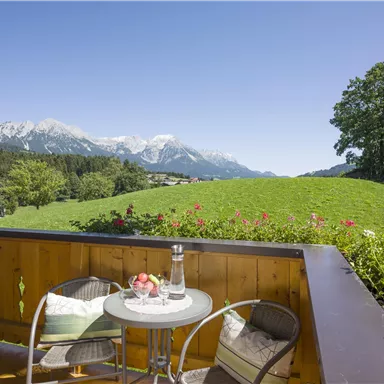 Ein gemütlicher Balkon mit einem kleinen Tisch und Stühlen. Im Hintergrund sind grüne Wiesen und schneebedeckte Berge unter einem klaren blauen Himmel zu sehen.