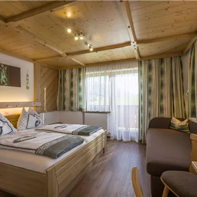 Ein gemütliches Hotelzimmer mit zwei Betten und einer Sitzecke. Große Fenster lassen viel Tageslicht herein und die Holzdecke sorgt für eine warme Atmosphäre.