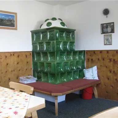 A room with a green tiled stove and wooden paneling. A round table with a colorful tablecloth and chairs can also be seen.