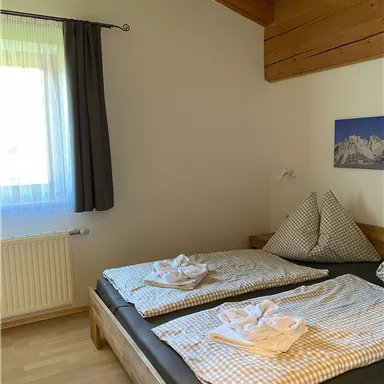 A cozy bedroom with two beds and wooden ceilings. The windows let in plenty of daylight, and the atmosphere is welcoming.