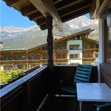 A balcony with a view of the mountains and a cozy seating area. The surroundings are characterized by wooden architecture and green plants.