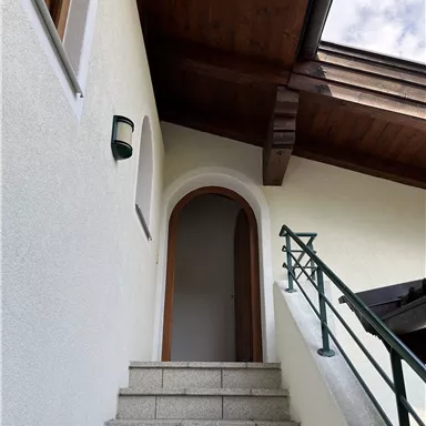 A staircase leads to an entrance under a wooden overhang. The walls are bright and the door is architecturally appealing.