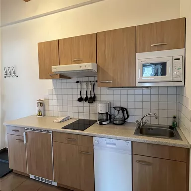 A modern kitchen with wooden fronts and bright tiles. It is equipped with a stove, microwave, coffee machine, and dishwasher.