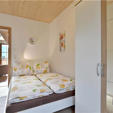 Ein gemütliches Schlafzimmer mit einem Doppelbett und Holzdecke. Im Hintergrund ist ein heller Wohnbereich mit einem Fenster und einem Stuhl zu sehen.