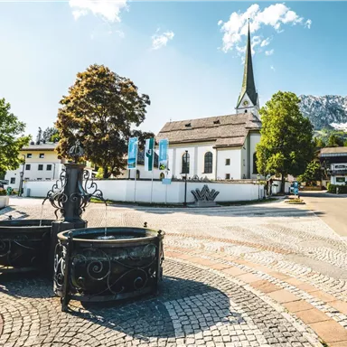 A picturesque village square with a church in the background and a clear sky. Surrounded by trees and historic fountains, the scene radiates tranquility and idyll.