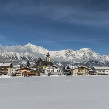 A snow-covered landscape with a small village and mountains in the background. The sky is clear and blue.