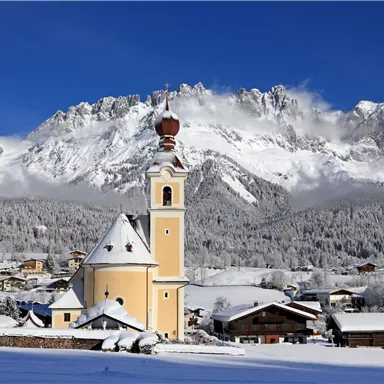 A picturesque mountain landscape with snow-covered peaks and a radiant blue sky. In the foreground stands a charming church with a distinctive tower.