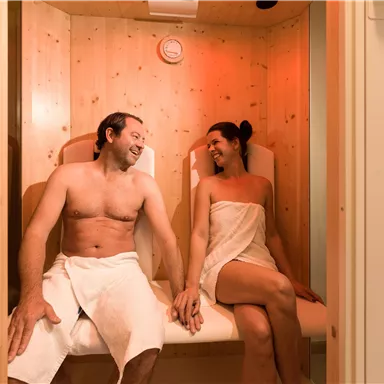 A couple sits relaxed in a sauna. They smile at each other and enjoy the warm wooden paneling.